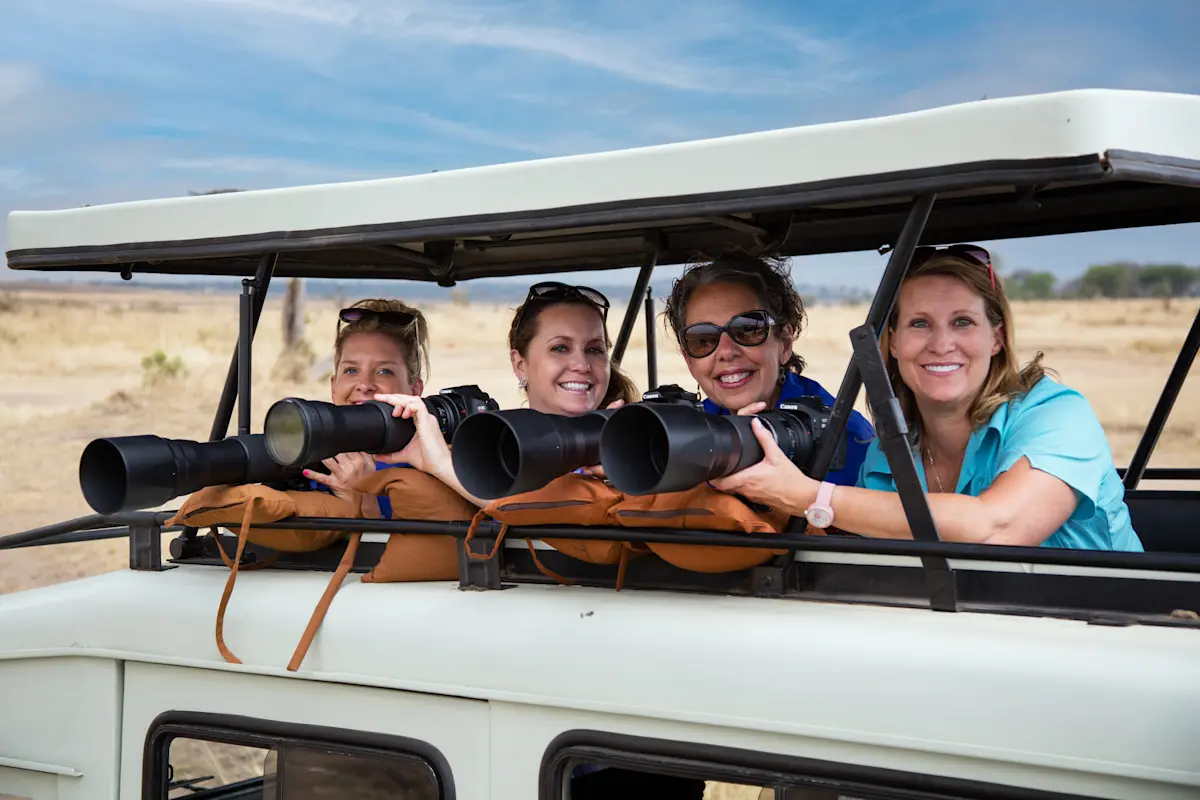 Four smiling guests with long camera lenses leaning out of an open-roof safari vehicle
