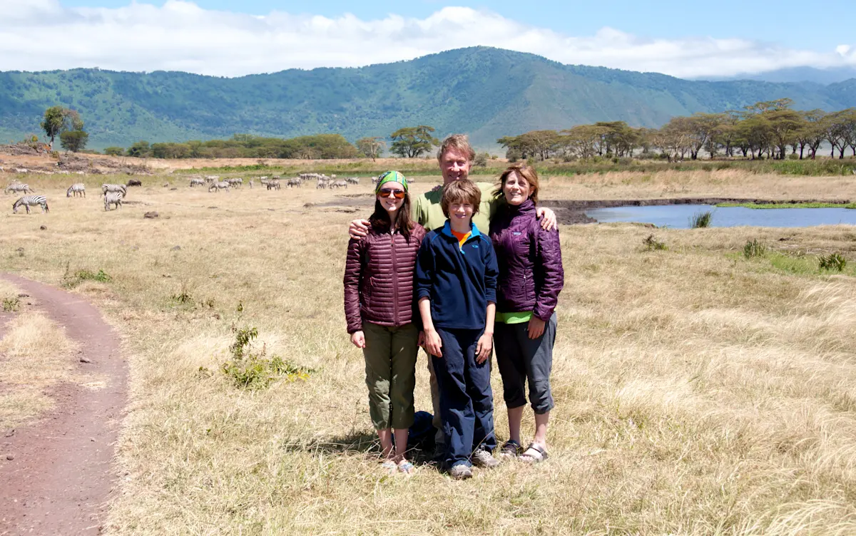Family of four posing on grassland with zebras and green hills behind