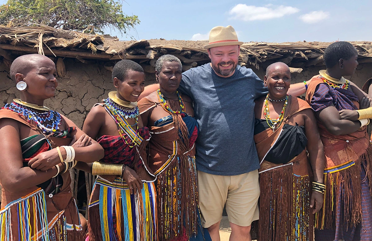 Guest posing with a group of women in beaded jewelry outside a mud hut