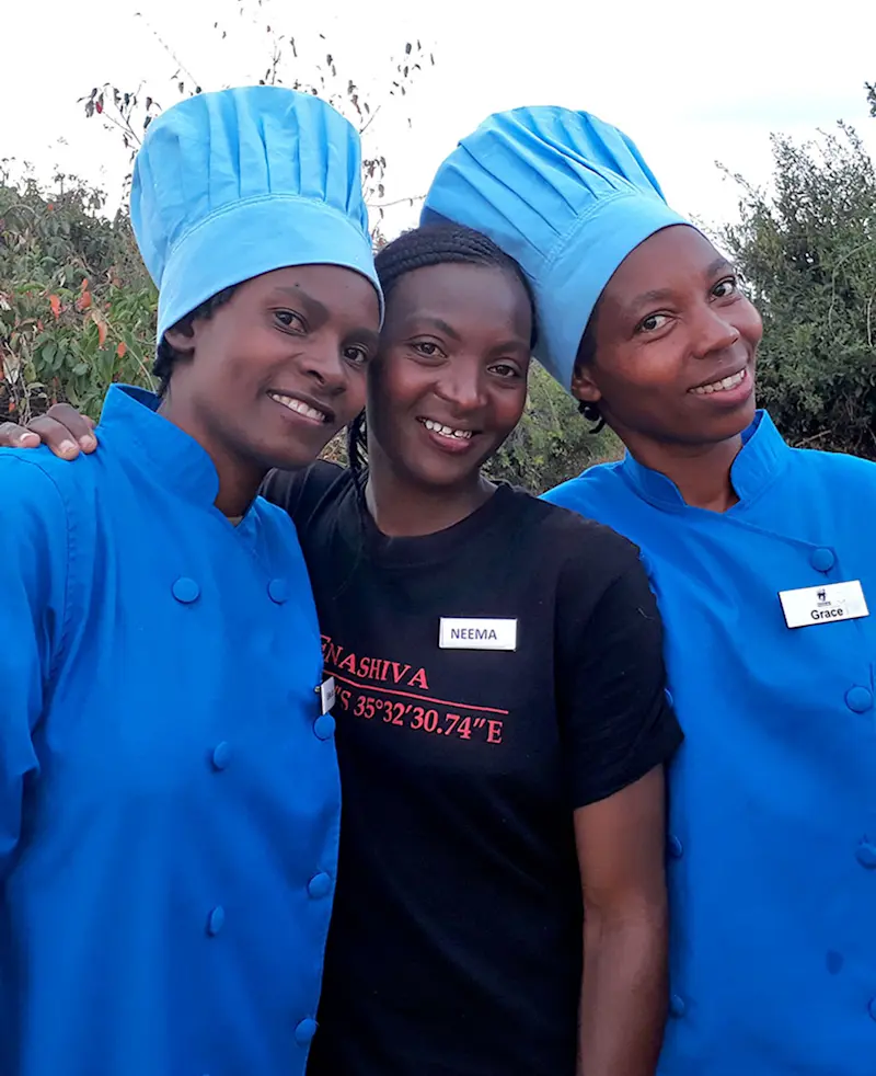 Three Nyumba camp staff women, two in blue chef uniforms, smiling together