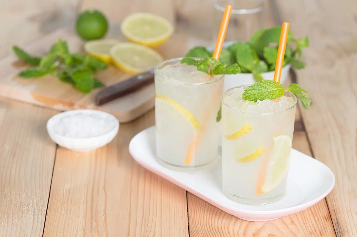 Two lemon and mint drinks with orange straws on a tray beside limes and lemons
