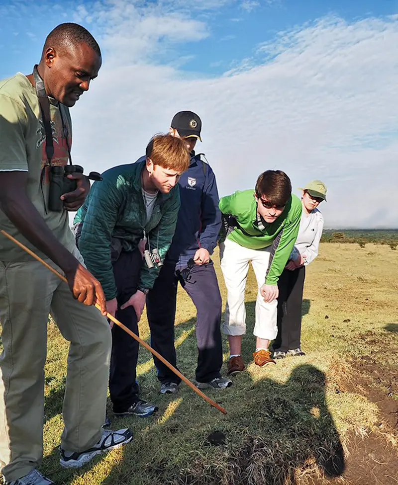 Guide pointing a stick at tracks on the ground as travelers lean in to look