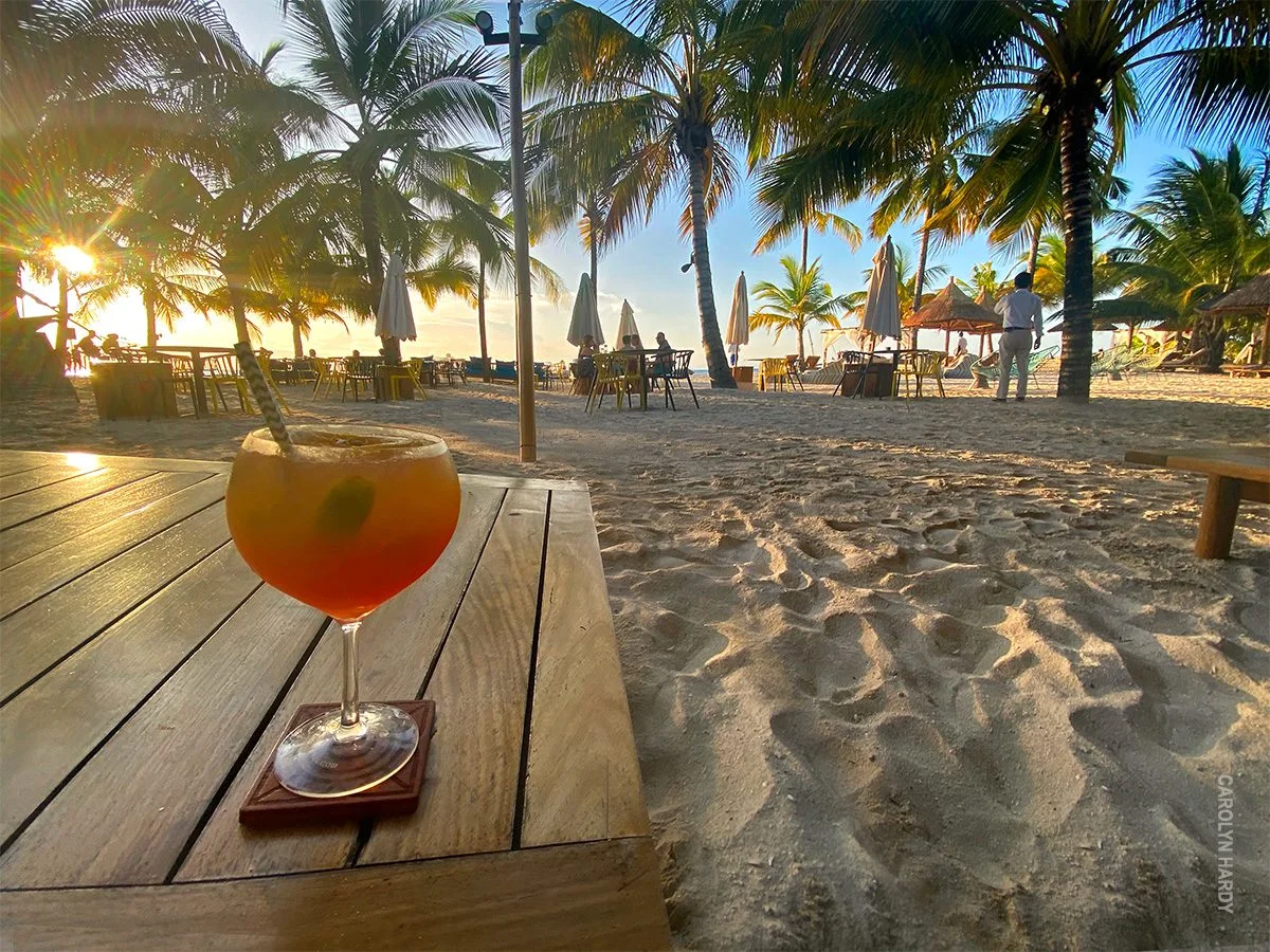 Cocktail on a wooden table on a sandy beach lined with palm trees at sunset