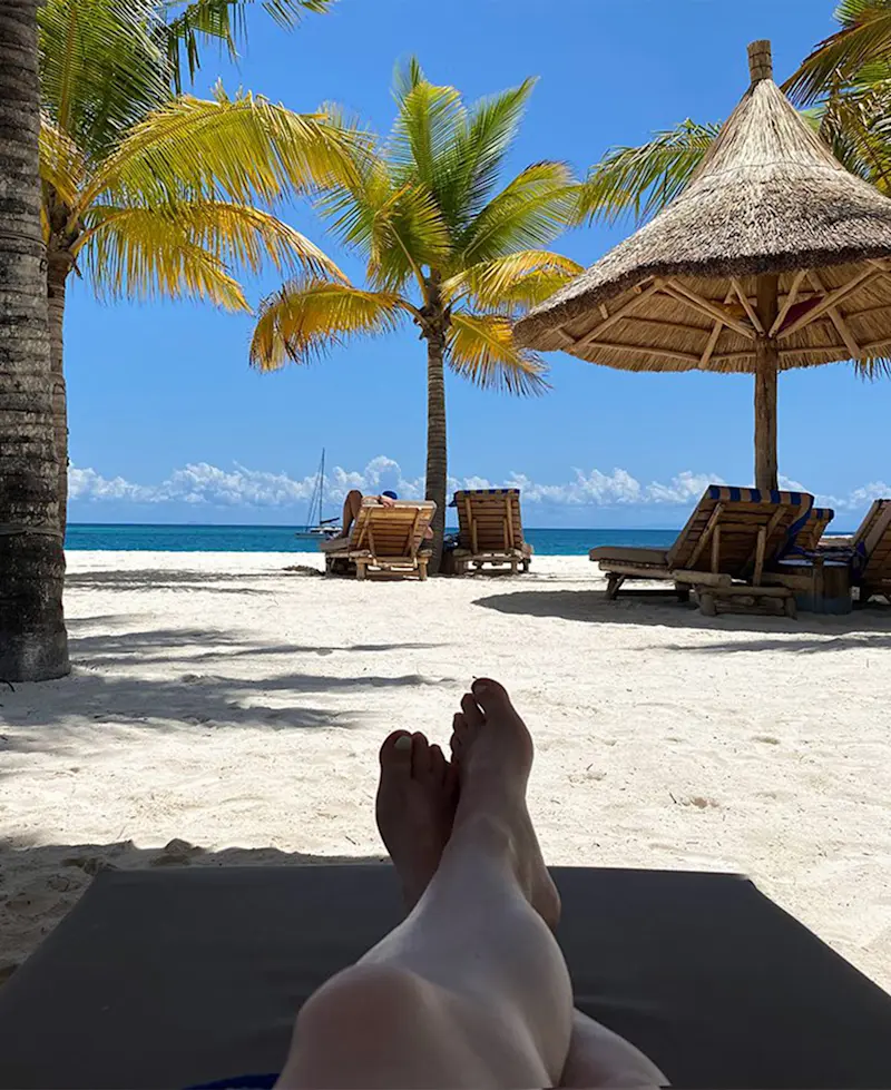 Feet up on a lounger facing a white sand Zanzibar beach with palms and thatched umbrellas