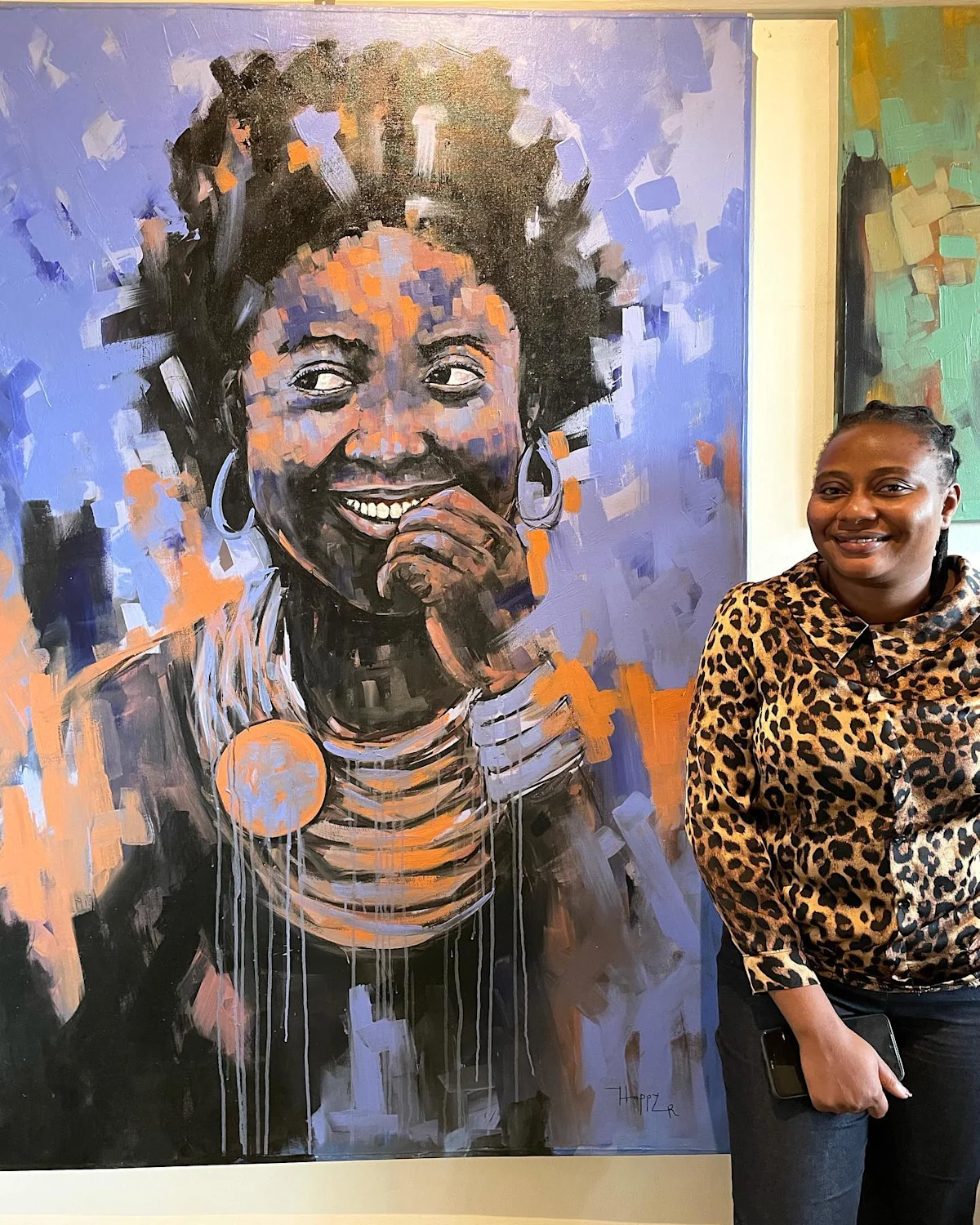 Woman standing beside a large painted portrait of a smiling woman in beaded necklaces