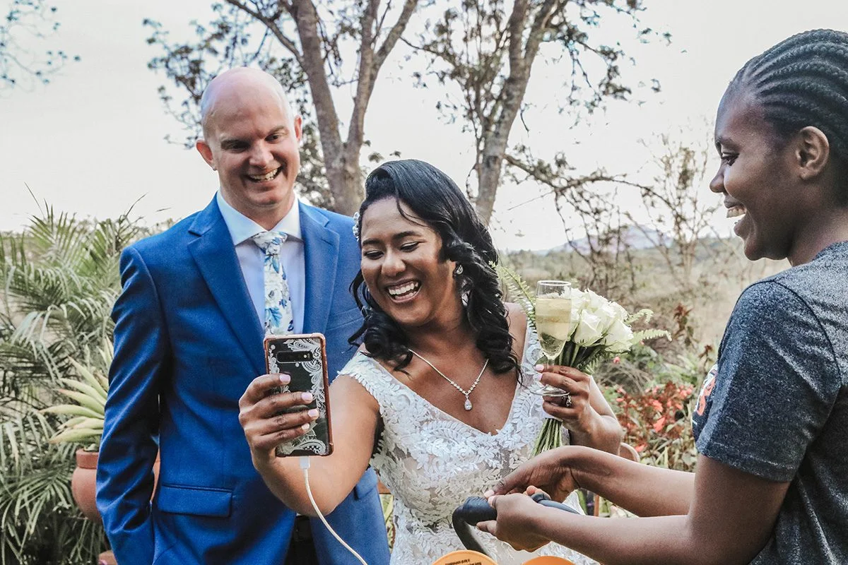 Laughing bride and groom holding a phone to greet wedding guests on a video call