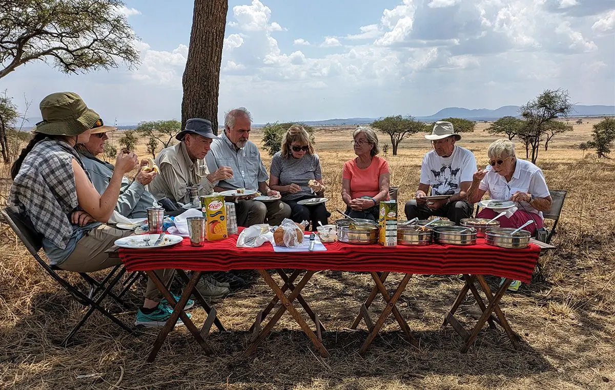 Guests eating a bush lunch at a long table under a tree in the Serengeti