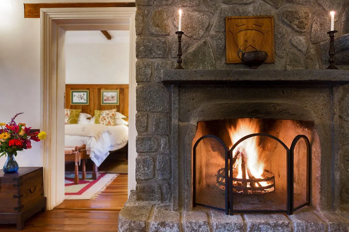 Fire burning in a stone fireplace with a bedroom visible through the doorway at Gibb's Farm