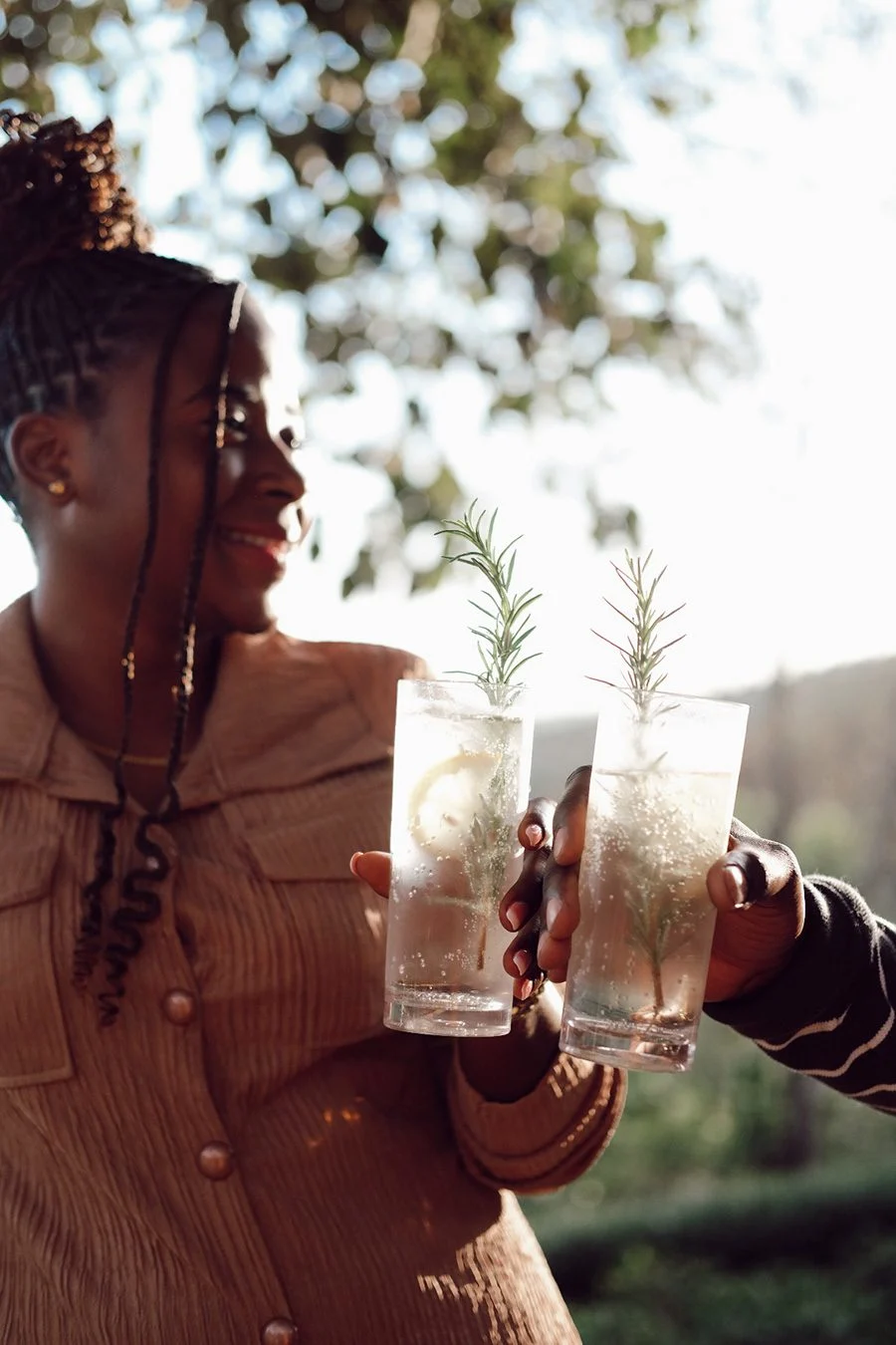 Two people toasting with sparkling drinks garnished with rosemary in evening sun