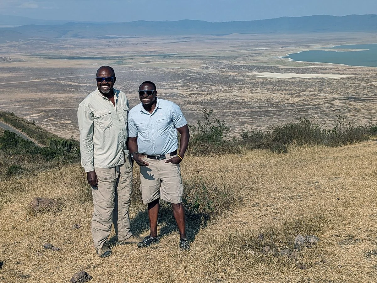Two safari guides standing on a crater rim above a wide crater floor and lake