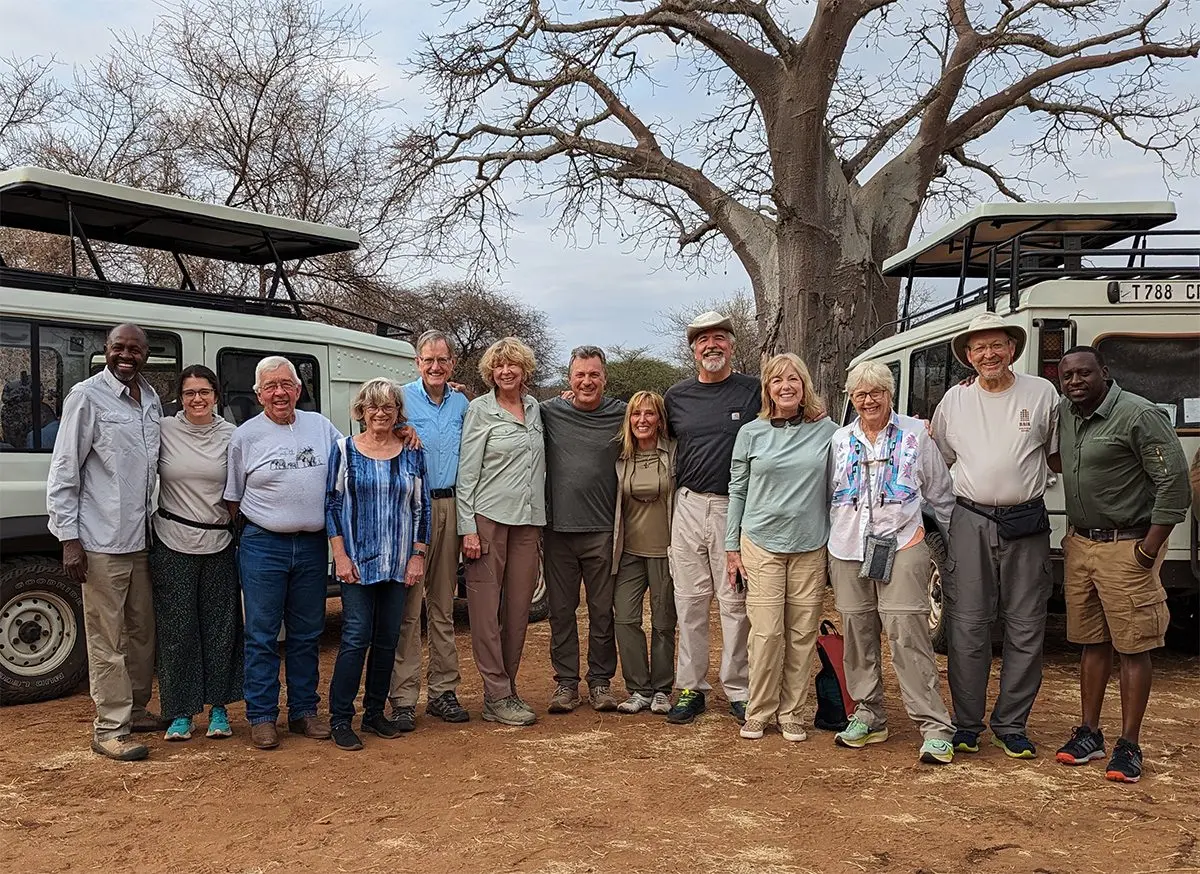 Large group of Thomson travelers and their guides posing by safari vehicles under a baobab