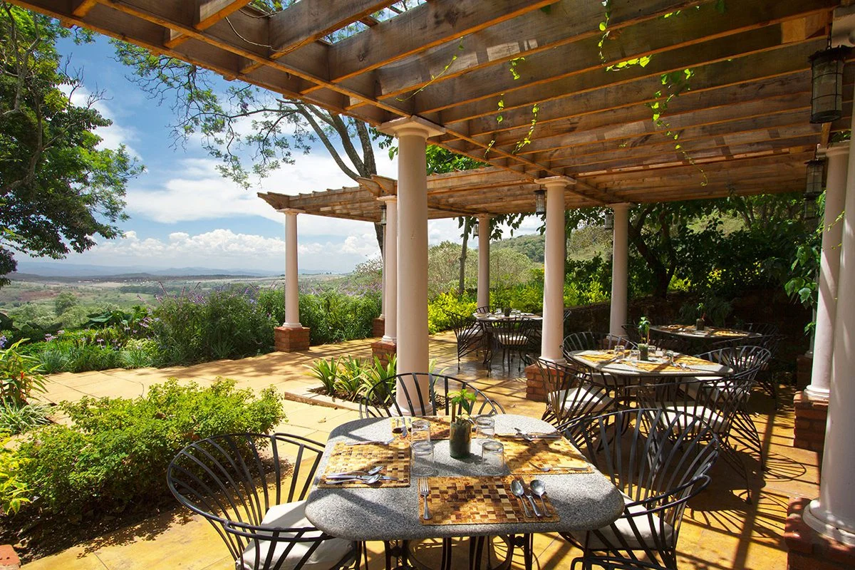 Outdoor dining terrace at Gibb's Farm under a wooden pergola with views of the hills