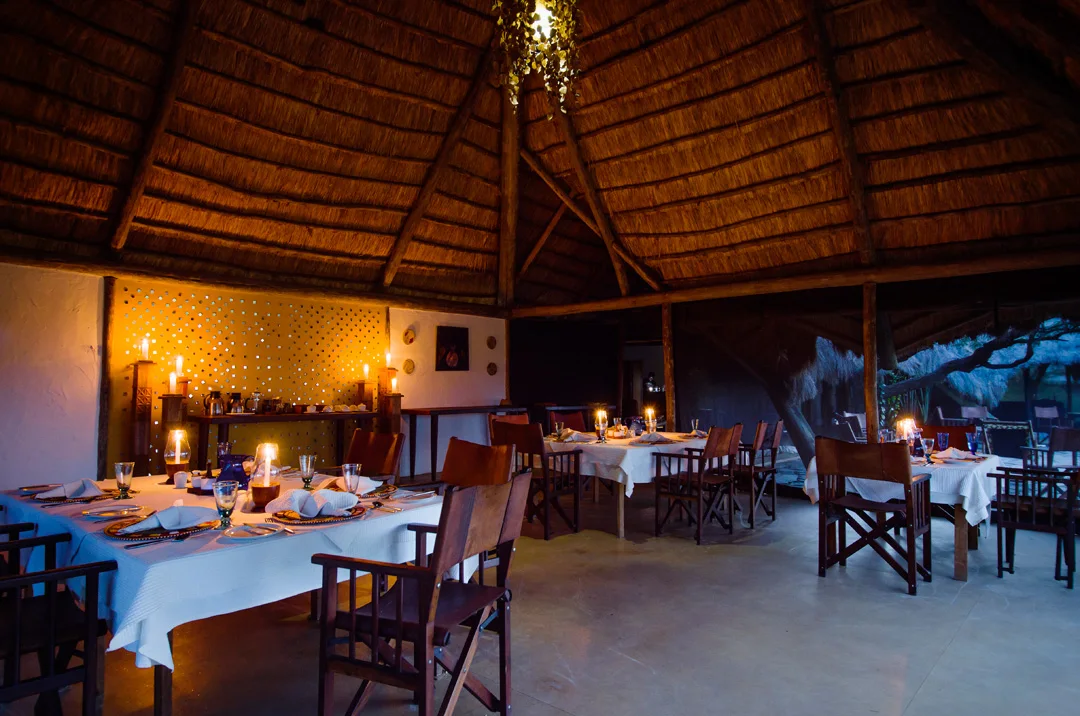 Candlelit tables set for dinner in a thatched-roof camp dining room