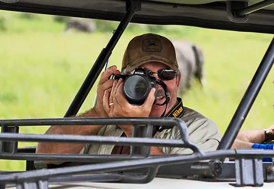 Man in a cap photographing with a Nikon camera from a safari vehicle