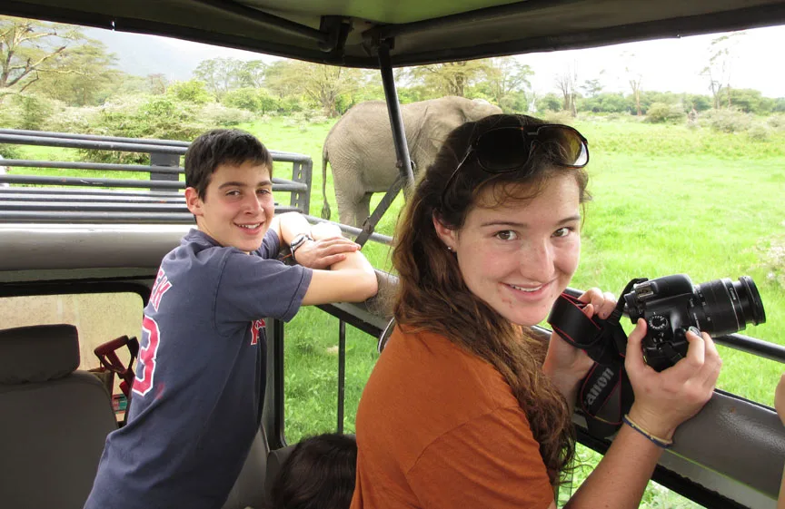 Two teens smiling in a safari vehicle with a camera as an elephant walks by