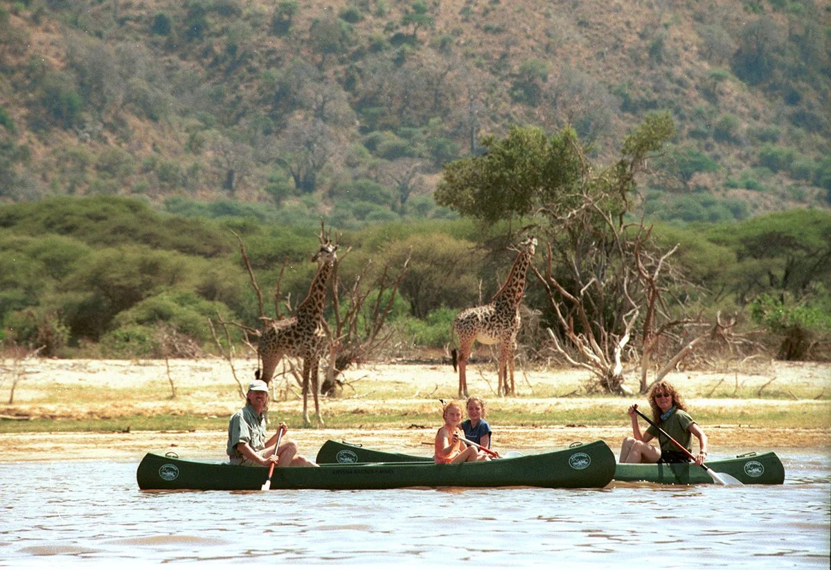 Family paddling canoes on Lake Manyara as giraffes stand on the shore