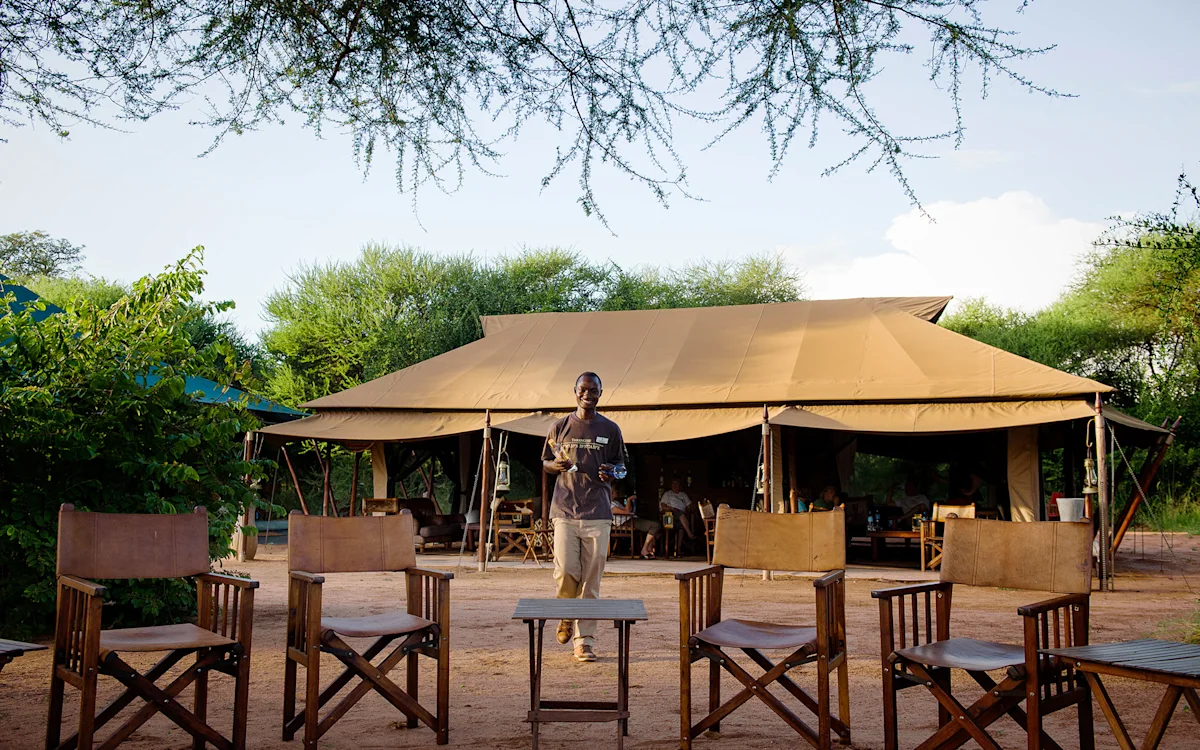Staff member carrying a tray past camp chairs outside the lounge tent at Tarangire Nyumba