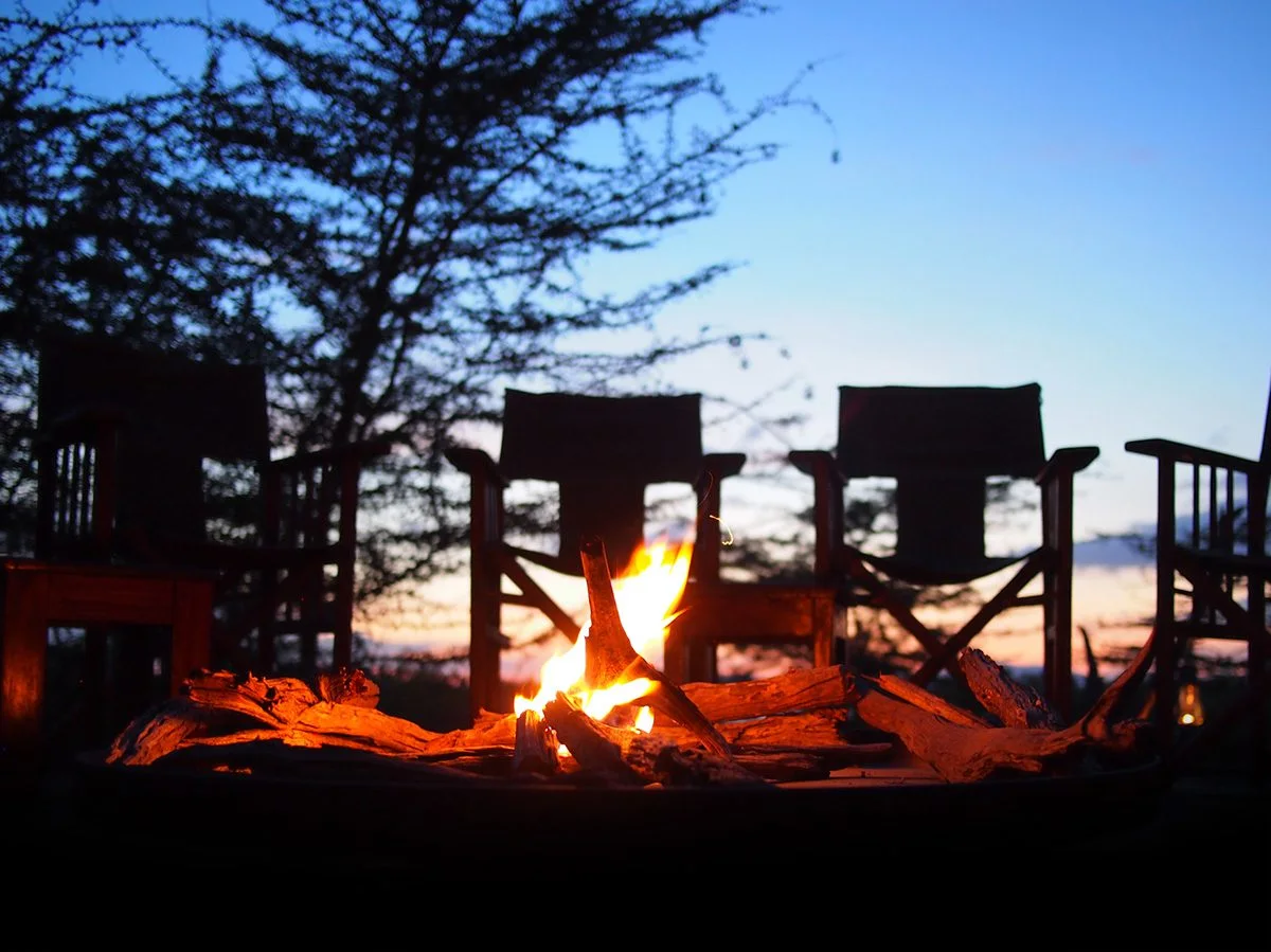 Campfire burning in front of empty safari chairs at dusk