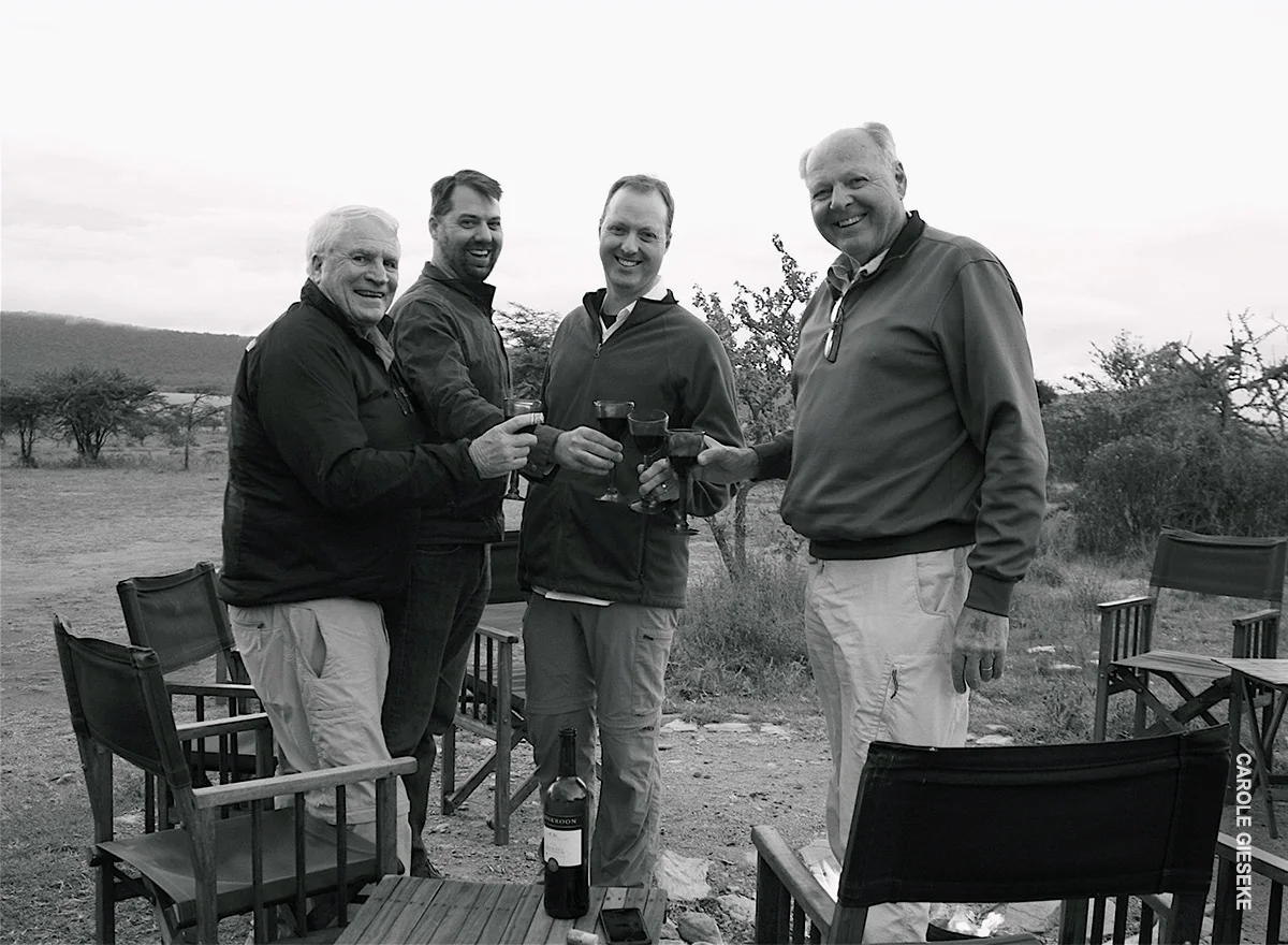 Black and white photo of four men toasting with wine around a camp fire pit