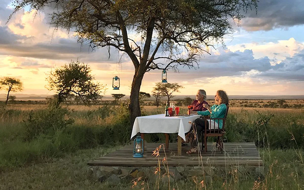 Rick Thomson and Judi Wineland at a table on a wooden deck beneath an acacia tree at sunset, Serengeti Nyumba