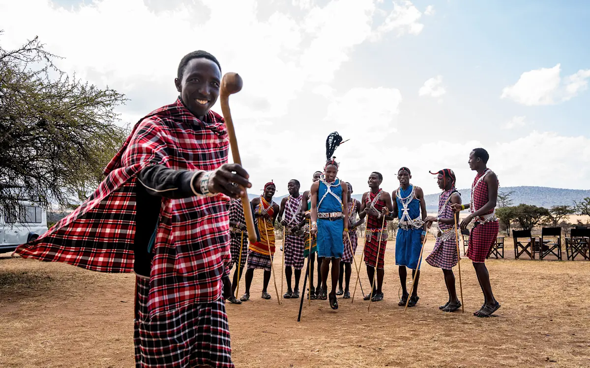 Smiling Maasai man in a red plaid shuka with a group of Maasai dancers behind him
