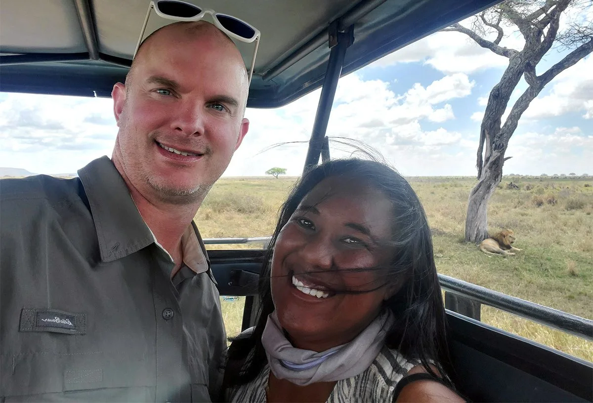Couple taking a selfie in a safari vehicle with a lion resting under a tree behind them