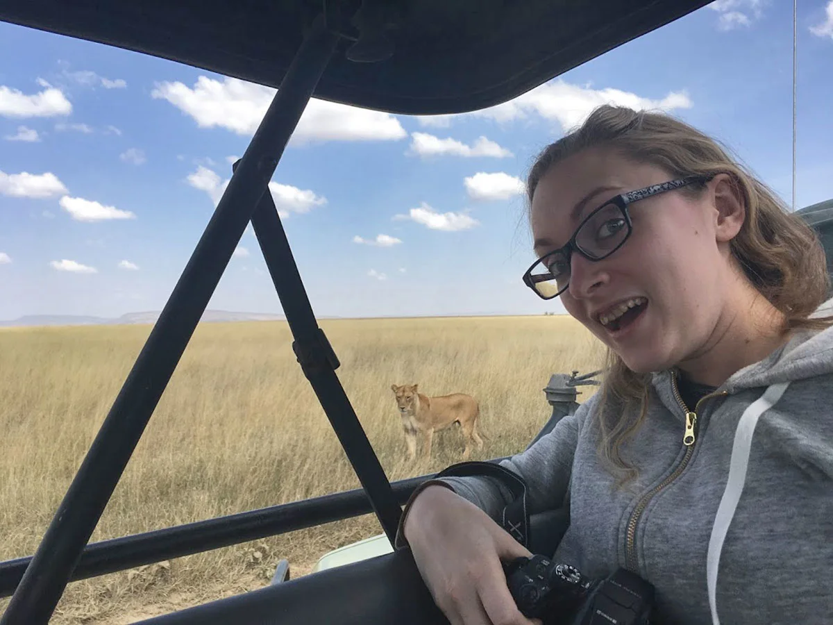 Smiling traveler with a camera in a safari vehicle as a lioness walks through the grass beyond