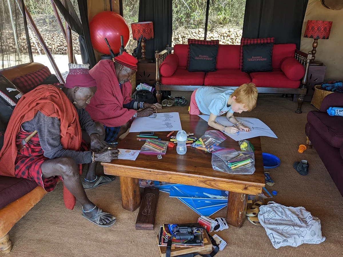 Maasai elders and a child coloring pictures together at a table in a lounge tent