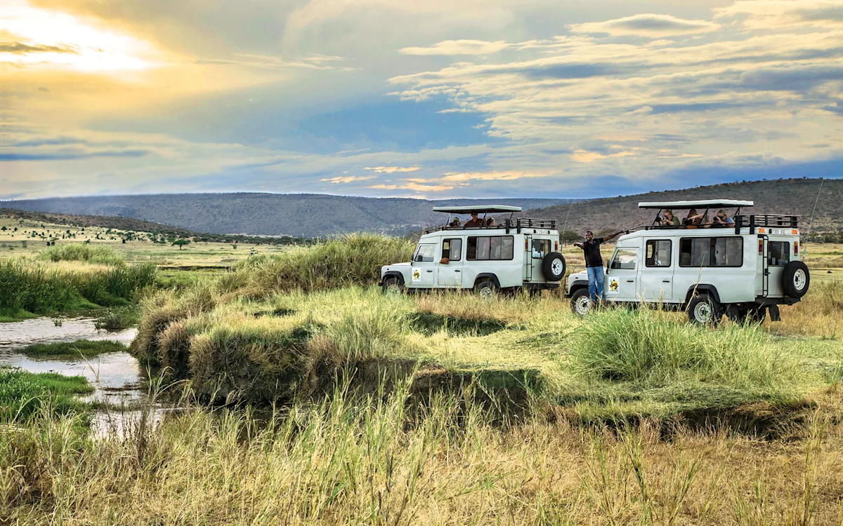 Safari vehicles parked by a stream on grassy plains as guests look out