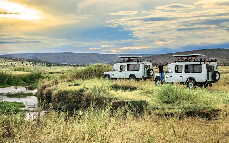 Safari vehicles parked by a stream on grassy plains as guests look out