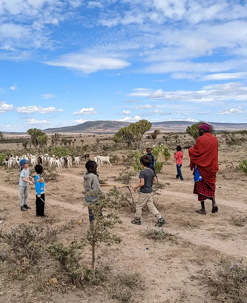 Children walking with a Maasai herder and a herd of goats across dry scrubland