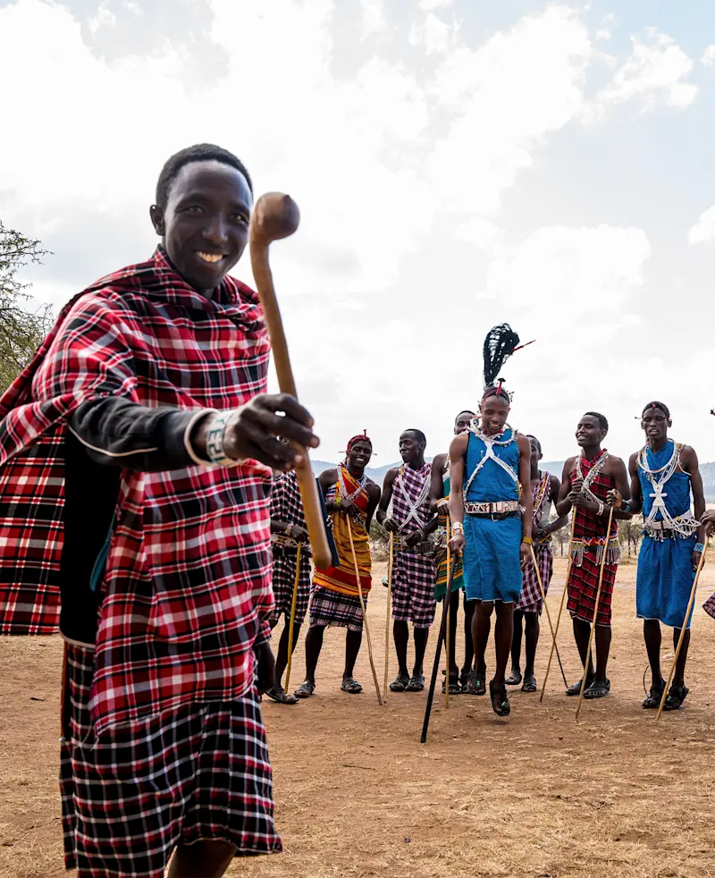 Smiling Maasai man in a red shuka as warriors behind him perform a jumping dance