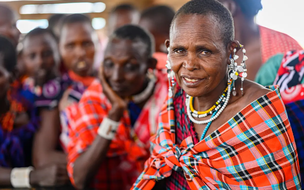 Maasai woman in a plaid shuka and beaded jewelry sitting with community members at a meeting