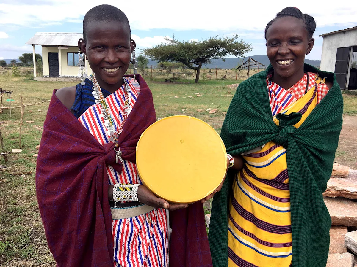 Two smiling Maasai women holding a round wheel of beeswax