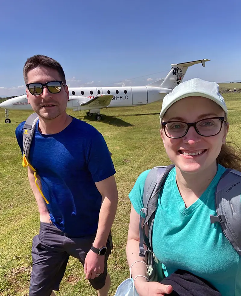 Smiling couple with backpacks on a grass airstrip beside a small plane
