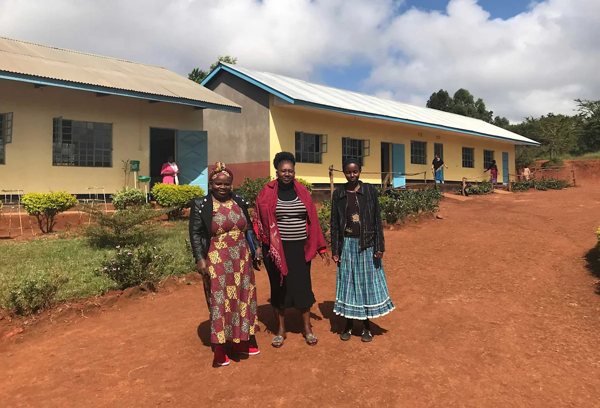 Three women standing in front of a row of school buildings on a red dirt path