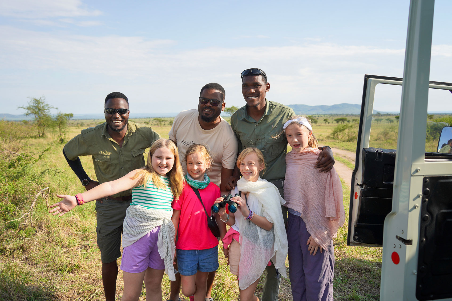 Three guides posing with four young girls beside a safari vehicle