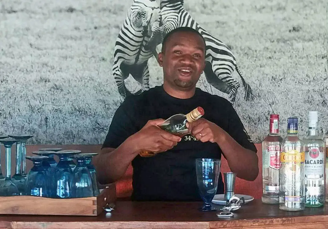 Smiling bartender holding a bottle at a camp bar with a zebra mural behind