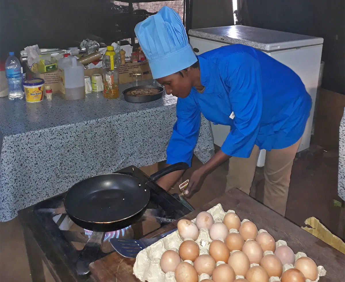 Chef in a blue uniform and hat cooking at a camp stove beside trays of eggs