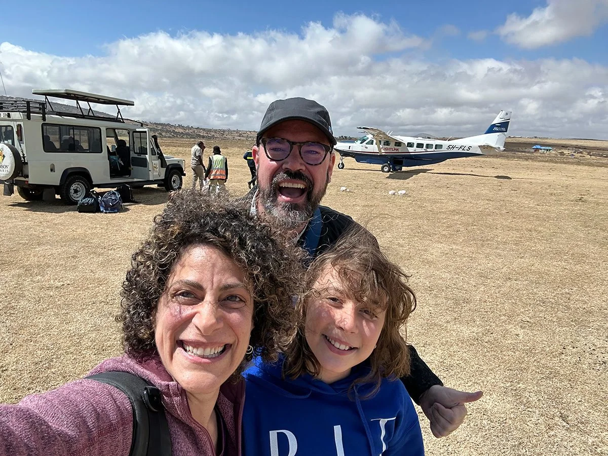 Smiling family taking a selfie on an airstrip with a safari vehicle and small plane behind