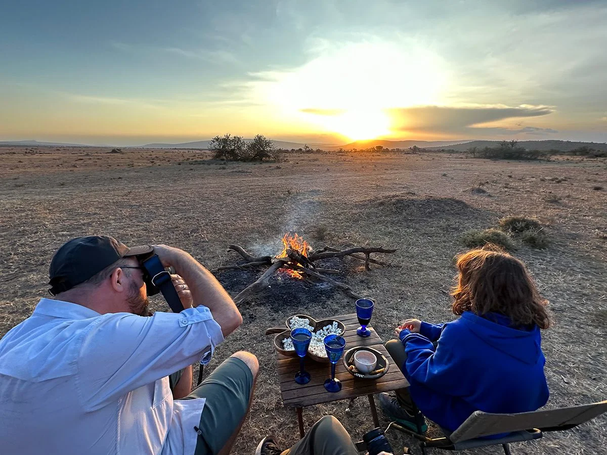 Two travelers enjoying drinks and snacks by a campfire as the sun sets over the plains