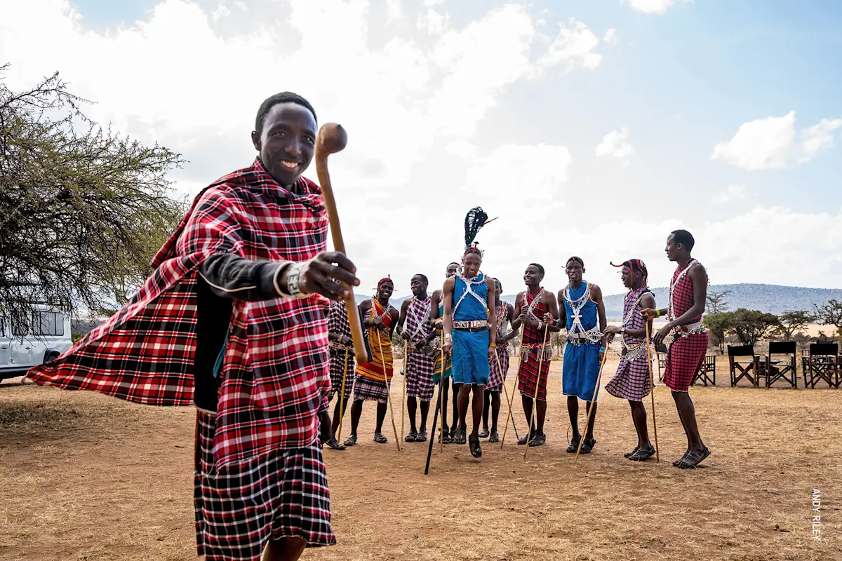 Maasai man in a red plaid shuka holding a club, with Maasai men dancing behind him