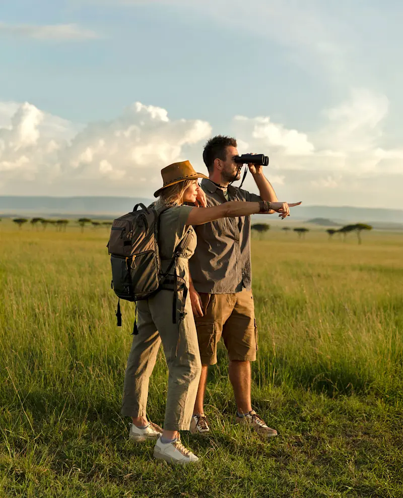 Couple on a walking safari, one looking through binoculars as the other points across the grassland
