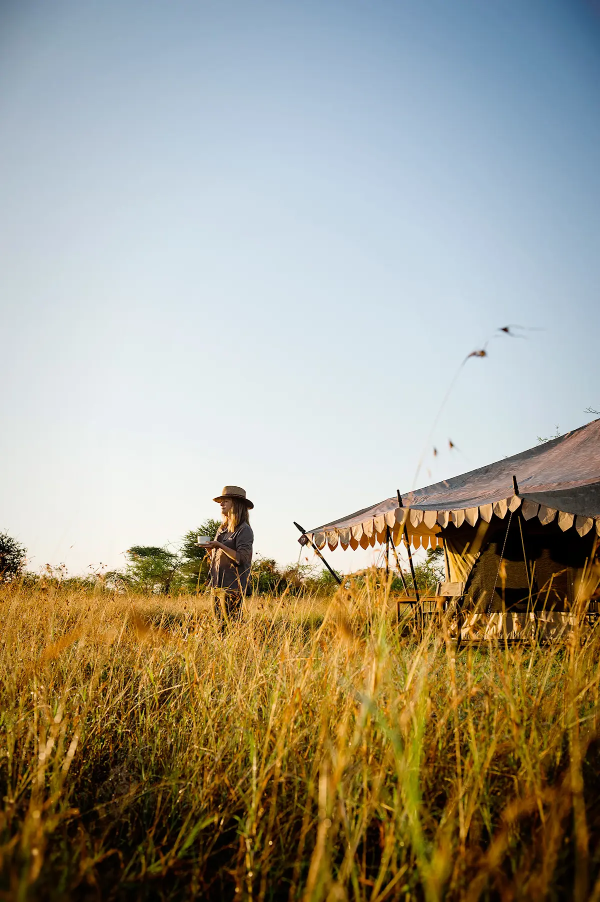 Woman with a coffee cup in tall grass beside a canvas tent at a Nyumba camp