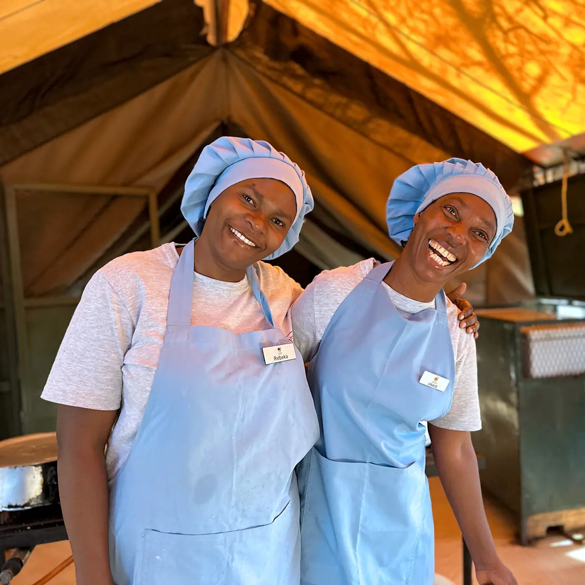 Two camp chefs in aprons and caps smiling inside a kitchen tent