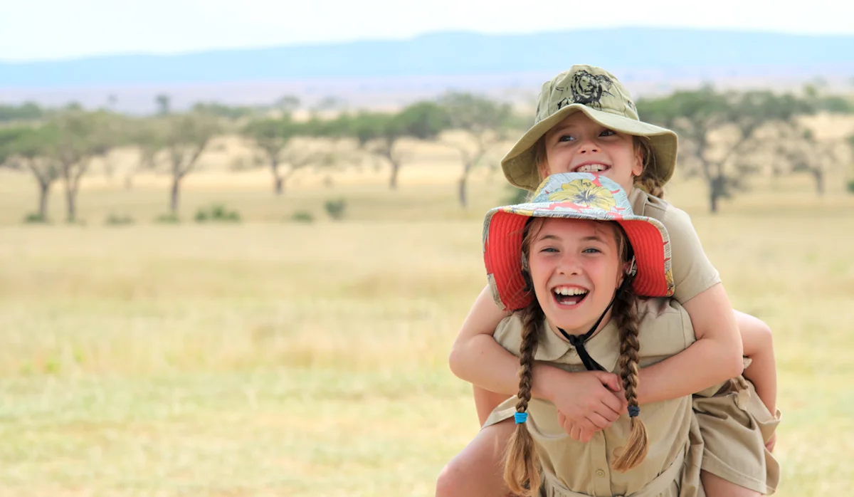 Two laughing girls in safari hats, one riding piggyback, on the savanna