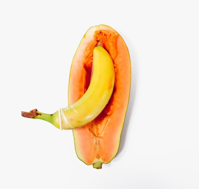 En banan med kondom placeret inde i en halv papaya på hvid baggrund.