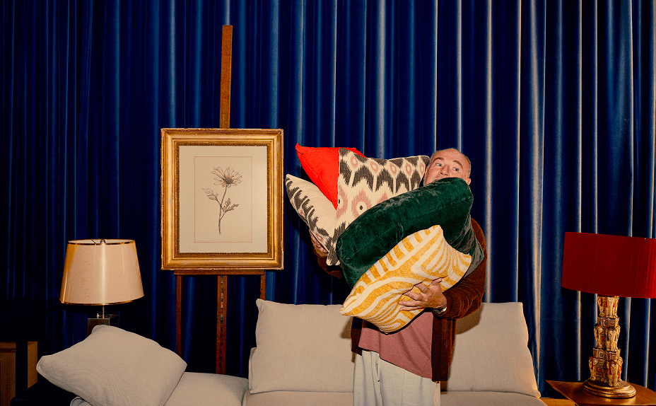 A person in a room with dark blue curtains in front of a cream sofa, holding too many small pillows