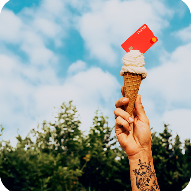 A tattooed hand holds up a vanilla ice cream cone with a bright coral Monzo bank card inserted into the top, set against a backdrop of green trees and a cloudy blue sky
