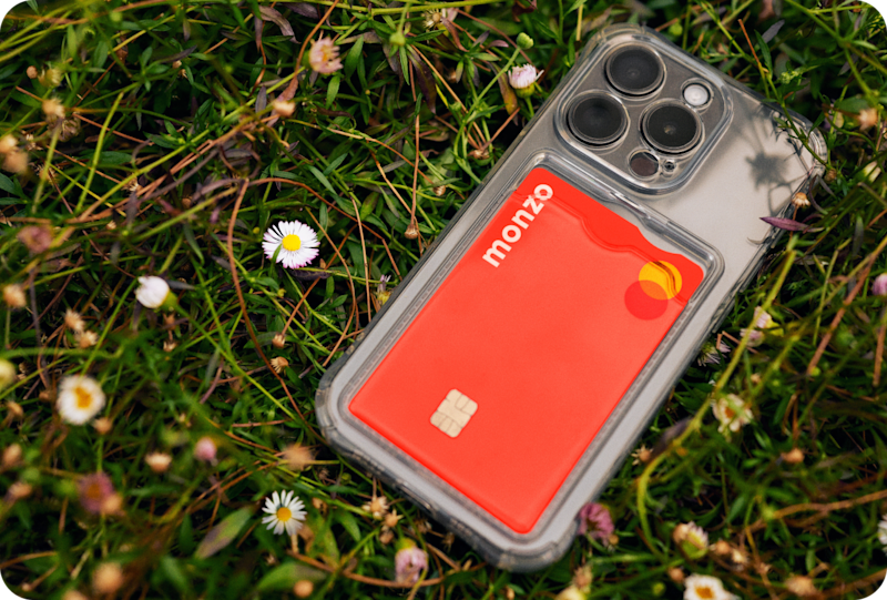Image on an iPhone on a grassy field, with the Monzo hot coral card in the back of the case.